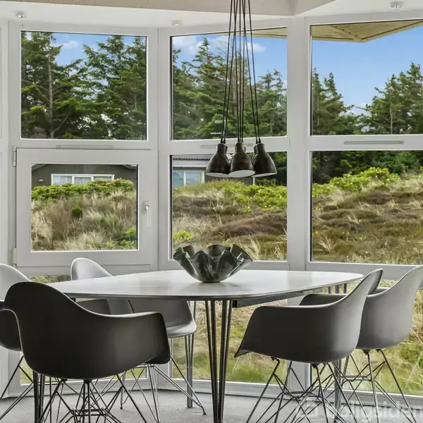 Spisebord med fem sorte stole står ved et stort vindue med udsigt til en skov. Over bordet hænger en moderne lampe, og udenfor ses et grønt landskab.