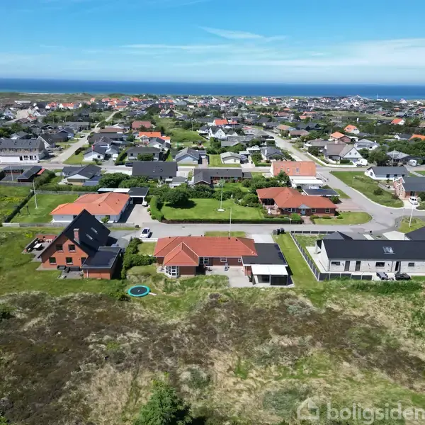 Huse ligger tæt i et grønt boligområde med kysten i baggrunden. Det åbne terræn foran viser græs og buske, mens blå himmel strækker sig over landskabet.