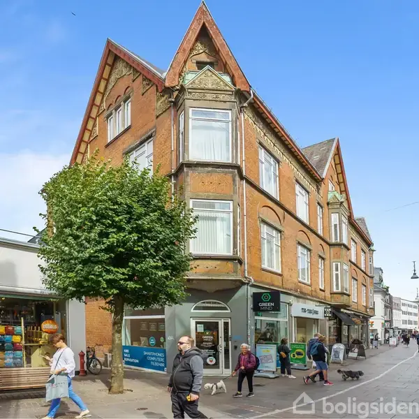 Et hjørnebygning i røde mursten med spidse gavle. Folk går på fortovet foran butikker i en travl gågade med blå himmel i baggrunden.