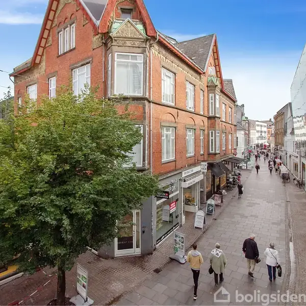 Bygning står langs en gågade, omgivet af butikker og fodgængere. En rød murstensbygning med dekorativ gavl pryder hjørnet. En stor grøn træ står ved siden af facaden.