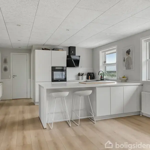 Køkken med hvid køkkenø og to barstole, placeret i et rummeligt værelse med lyse trægulve og to vinduer, der giver naturligt lys. Radiator og maleri på væggen.