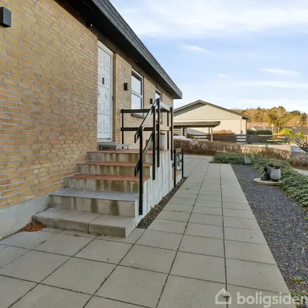 En bygning med murstensvægge står ved siden af en flisebelagt sti. Trapper fører til en hvid dør, omgivet af hække og græs i et boligområde med garager i baggrunden.