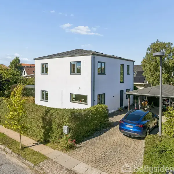 Et hvidt, to-etagers hus står stille på en solrig dag omgivet af hække. En blå bil er parkeret på grusindkørslen ved siden af huset, i en rolig beboelsesgade.
