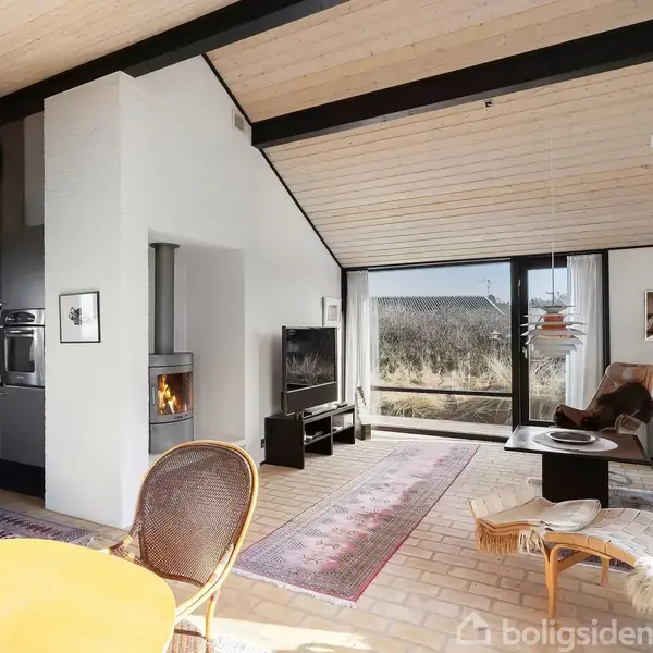 Stuen har et moderne design med trægulv og store vinduer. En brændeovn er tændt, og der står møbler som stole og sofabord. Udenfor ses buske gennem vinduet.
