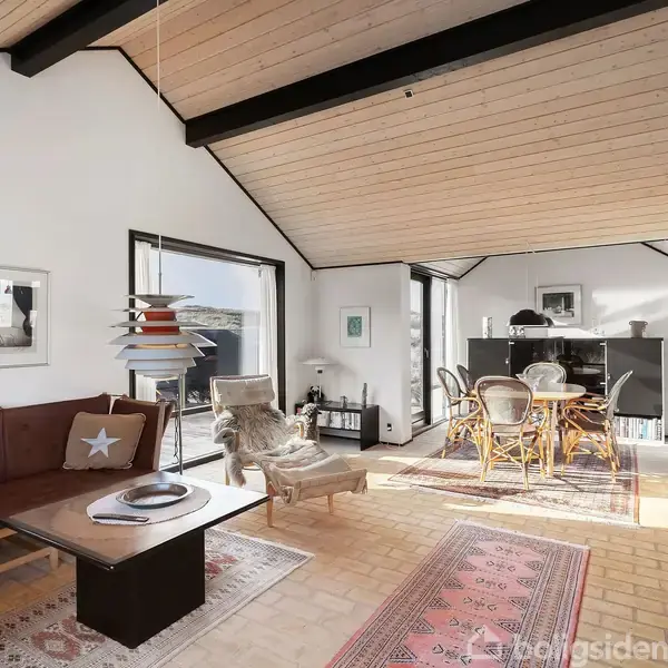 Stue med brun sofa og sofabord ved siden af et stort vindue. Træloft med hængelampe. Spisebord med stole i baggrunden. Lyse vægge og tæpper på gulvet.
