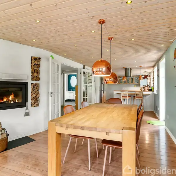 Stort spisebord med stole står i et lyst rum. En pejs med bunke brænde ved siden af. Kobberlamper hænger over bordet. Verdenskortmaleri på væggen til højre.