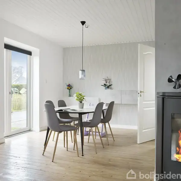 Et moderne spisebord med fire stole står i et lyst rum med store vinduer. En brændeovn til højre har ild i. En åben dør leder til et tilstødende værelse.