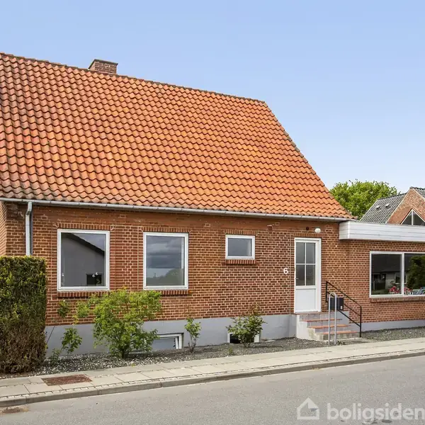 Et murstenshus med rødbrunt tegltag står ved en vej. Huset har en lille trappe ved indgangsdøren og er omgivet af lave buske og naboer.