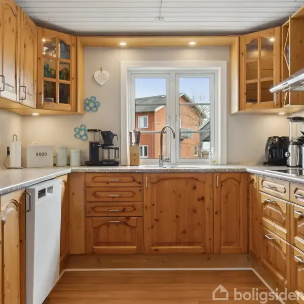 Køkken med træskabe og bordplader af granit. Køleskab og opvaskemaskine står til venstre. Kaffemaskine og knivblok på bordet. Vinduet afslører udsigt til et murstenshus.