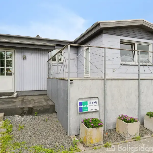 Grå hus med hvid dør har en terrasse foran. Skilte ved terrassen viser "Til salg" fra Nybolig. Blomsterkrukker står nederst i billedet. Fliser og grus danner indgangspartiet.