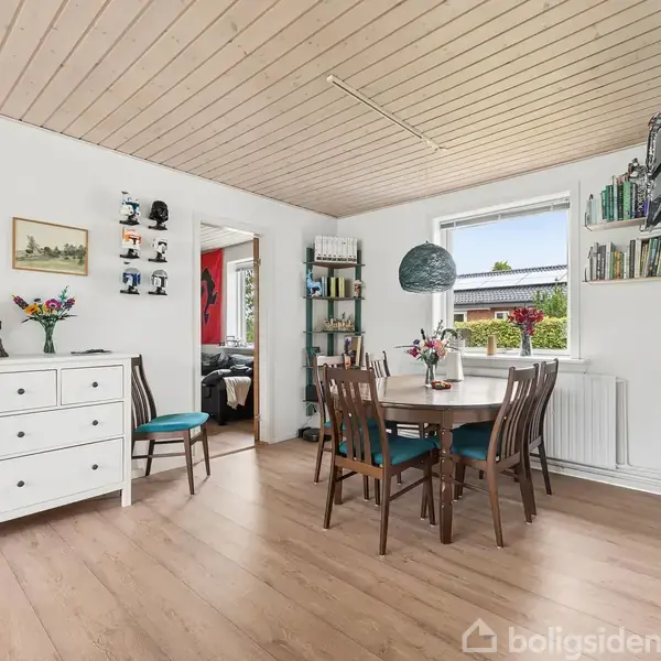 En spisestue med rundt spisebord og fire stole. En kommode med dekorationer står til venstre. Rummet har lysindfald fra et vindue og billeder på væggene.