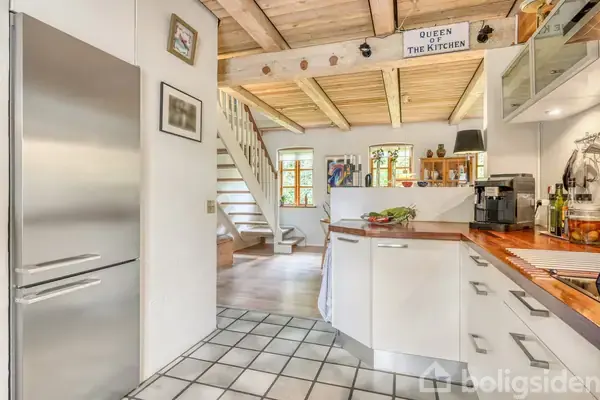 Et rustikt køkken med hvide skabe og en træbordplade. En skilt med teksten "QUEEN OF THE KITCHEN" hænger over indgangen til stuen, som leder til en trappe.