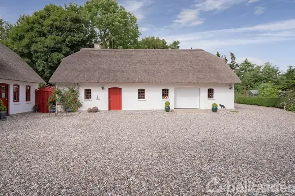 Hus med stråtag og hvid facade, omgivet af grusbelagt gårdsplads og haveplanter, står i landlige omgivelser med træer i baggrunden under en blå himmel.