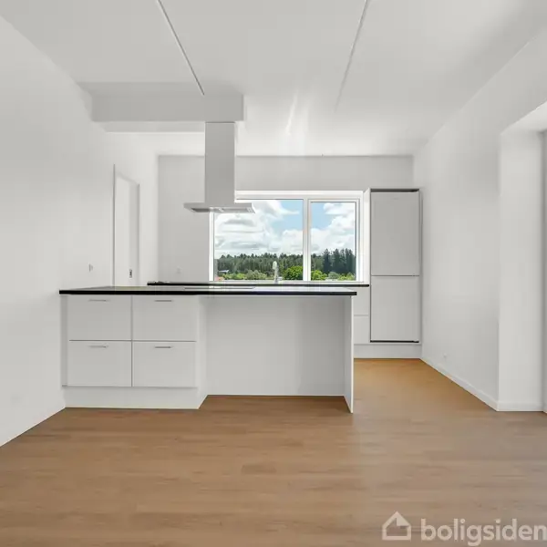 Et lyst køkken med hvide skabe og mørk bordplade. Køkkenet vender mod store vinduer, der åbner ud til en terrasse med udsigt over grønt landskab.