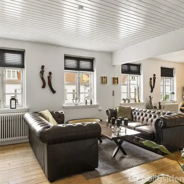 En stue med mørke lædersofaer står ved et sofabord. Væggene prydes af afrikansk-inspirerede dekorationer. Lyset strømmer ind gennem vinduer med sorte persienner. Trægulv og hvide vægge.