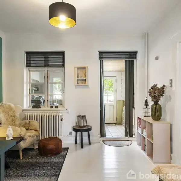 Lædersofaer og sofabord står i en lys stue med bræddegulve og hvidt loft. Væggene har kunstværker, og vinduerne har sorte rullegardiner, der slipper dagslyset ind.