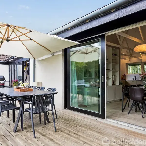 Udendørs terrasse med træbord og sorte stole under en parasol. Bagved åbner en skydedør til et moderne køkken med spiseplads. Trægulv omkranser området med grøn udsigt.