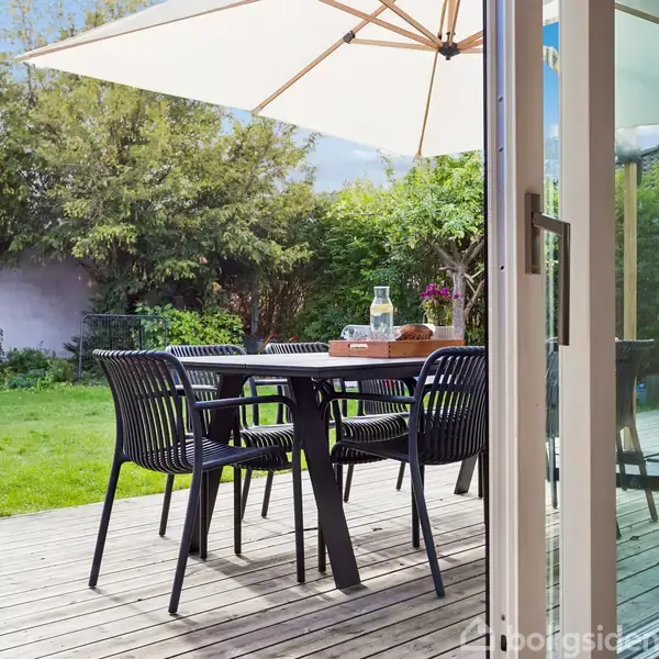 En udendørs terrasse med et bord og fire stole står under en stor parasol. En kande og et glas er på bordet. Haven i baggrunden har grønne træer og græs.