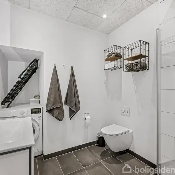Badeværelse med vaskemaskine ved siden af en håndvask med spejl. Grå håndklæder hænger på væggen. Toilettet er ved siden af bruser med glasdør. Hylder indeholder håndklæder og kurve.