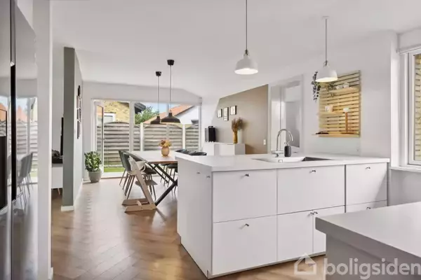 En moderne, lys køkkenalrum med en stor, hvid køkkenø i centrum. Ved siden af er et spiseområde med borde og stole. Store vinduer giver udsigt til haven udenfor.