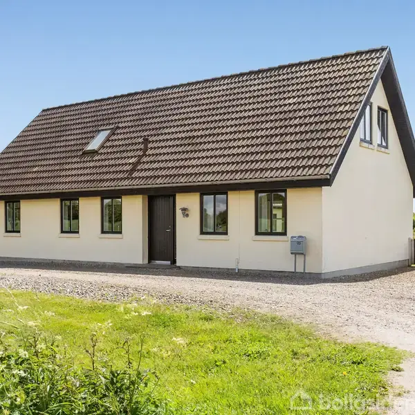 Et en-etagers hus med skråt tegltag står på en åbent område med grusindkørsel og grønne plæner omkring. Husets facade har flere vinduer og en enkelt dør.