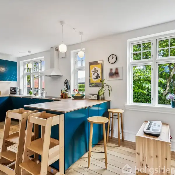 Køkken med blåt køkkenskab, træstole ved kogeø, og moderne indretning. Hvide vægge og store vinduer med udsigt til frodig have. Klokke og kunstværk hænger på væggen.