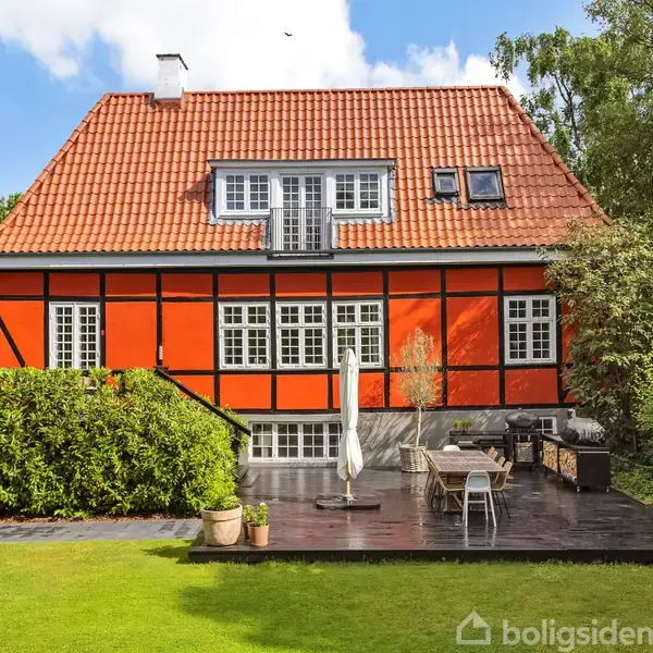Et rødt bindingsværkshus med rødt tegltag. Foran huset ses en terrasse med havemøbler og parasol. Græsplæne og træer omgiver huset i en frodig have.