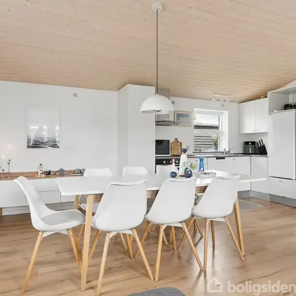 Spisebord med hvide stole er centreret i et lyst køkken-alrum. Køkkenet har hvide skabe og stålkomfur. Trægulve og hvidmalede vægge skaber en moderne og minimalistisk atmosfære.