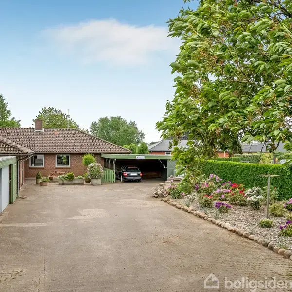 En garageport står åben ved et murstenshus med have omgivet af træer og blomsterbede. En bil er parkeret i carporten. En bred indkørsel fører til huset.