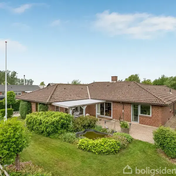 Et enfamiliehus med rødt tegltag står i en velplejet have med græsplæne og buske. Huset har en terrasse og et lille havebassin. Omgivet af stakitter og hække.