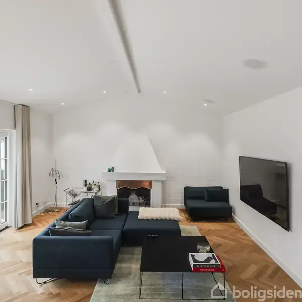 En moderne stue med en stor, mørkeblå hjørnesofa vendt mod en fladskærm på væggen. Spisebord ved glasdøre til altan. Lys trægulv og hvid pejs skaber en lys atmosfære.