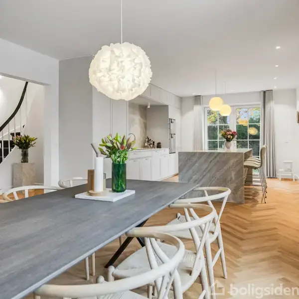 Langt bord med lyse træstole står i et åbent køkken-alrum. En vase med blomster pryder bordet. Moderne lamper hænger fra loftet. Trægulv og lyse vægge omkranser rummet.