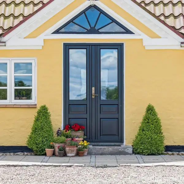 Dobbelt sort dør står mellem to vinduer på en gul væg. Foran døren er krukker med blomster og små buske, der skaber en indbydende atmosfære.