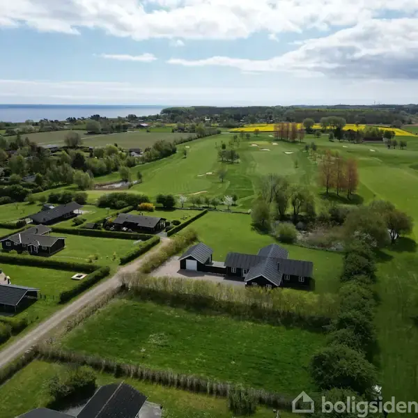 Huse ligger spredt langs veje i et grønt landskab med marker og en golfbane. I baggrunden ses havet under en delvist overskyet himmel.
