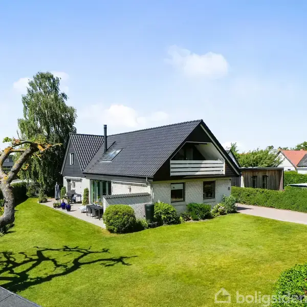Hus med mørkt tag og hvid facade står i en velplejet have. Træer og buske omgiver huset, og en indkørsel fører til fire skraldespande langs en hæk.