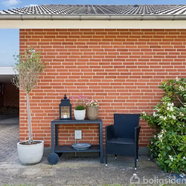 En sort stol står ved siden af et lille bord med lanterne og plantekrukke. Alt er placeret ved en murstensvæg med en hæk til højre og en carport til venstre.