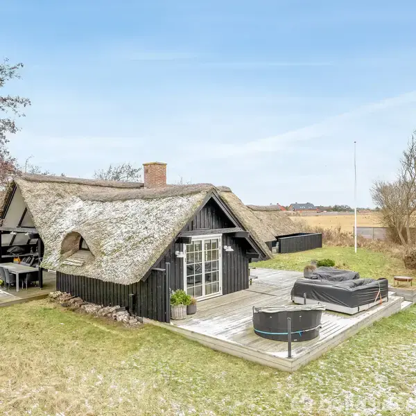 Et stråtækt hus står på en græsplæne med en træterrasse. Terrassen har udendørs møbler til afslapning. Der er nøgne træer og åbent landskab i baggrunden.