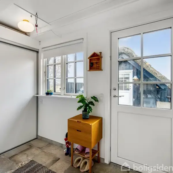 En lys entré indeholder en hvid dør med glasruder. Et lille bord med en plante står i hjørnet. Der er også et vindue og en skydedør.