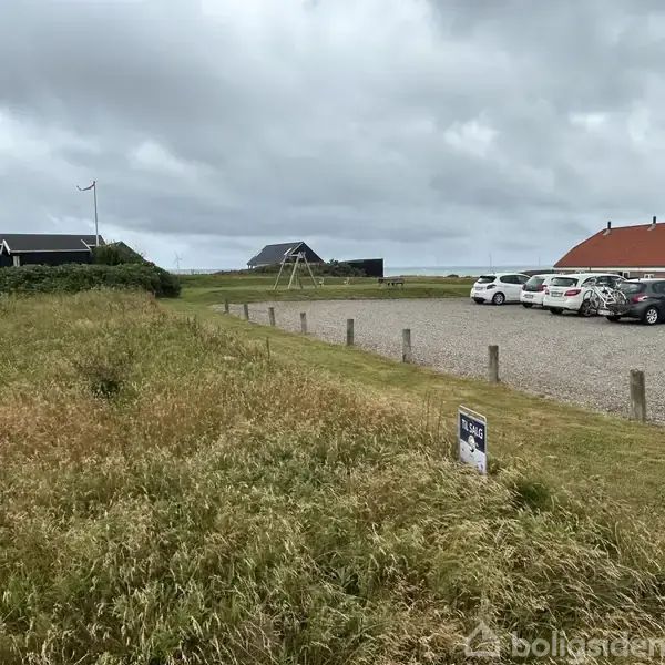 Græsareal med skiltet "Til salg" foran en parkeringsplads med biler. Omkranset af lave hegn og huse i baggrunden under en overskyet himmel.
