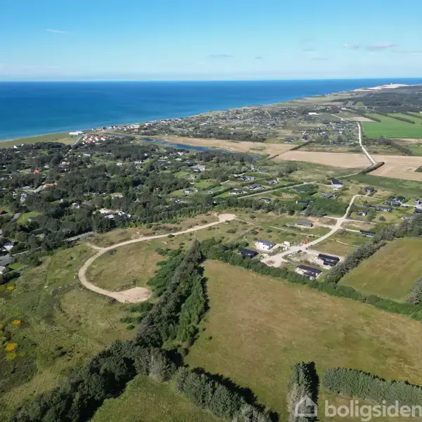Luftfoto viser et landskab med huse og marker, der strækker sig mod en blå kystlinje. Området er præget af bølgende grøn natur og spredte bebyggelser langs havet.