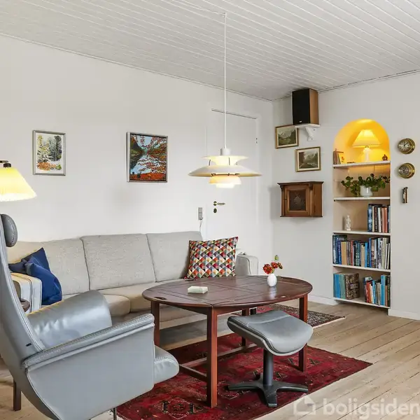 Grå lænestol og sofabord med vase i stue med lys sofa, reoler med bøger, lamper og nogle vægkunstværker. Rummets lysindfald skaber en hyggelig atmosfære.