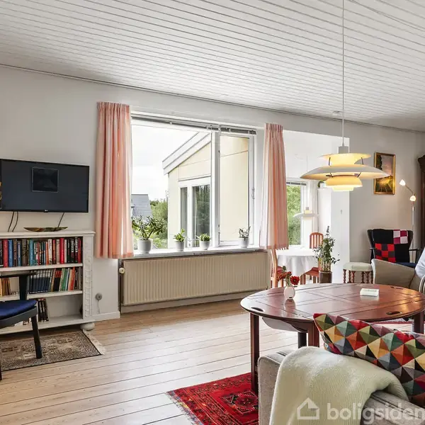 Stue med træbord og lænestol, omgivet af bøger på reol. Fjernsyn hænger på væggen. Store vinduer med gardiner giver lysindfald, og indretningen er hyggelig med tæpper og planter.