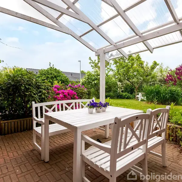 Hvidt træbord og stole står under en pergola af glas. Blomster i potter dekorerer bordet. Haven er frodig med buske og træer.