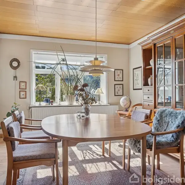 Et rundt spisebord er omgivet af træstole med pelsbetræk; en dækket glaslampe hænger over bordet. Rummet har store vinduer, der viser en have, og en stor træreol.