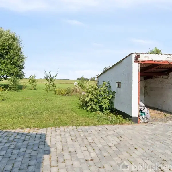 En åben garage uden port står på en brolagt gårdsplads. Baggrunden er en åben grøn mark med enkelte buske og træer under en blå himmel.