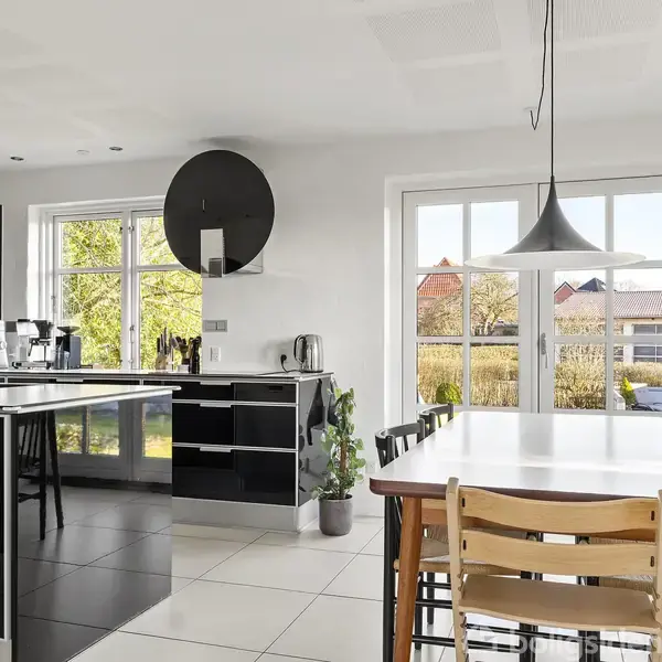 Køkken med sort skab, sort køkkenø med vandhane, placeret i moderne rum med store vinduer. Hvidt bord omgivet af stole i lyst, luftigt lokale med udsigt til have.