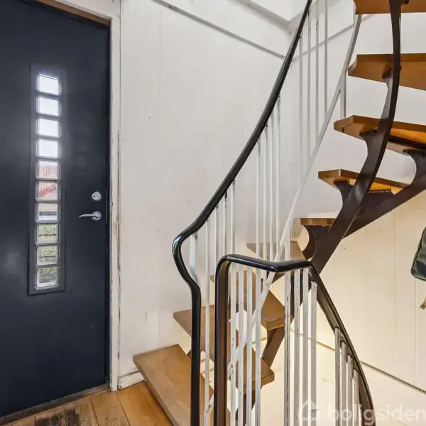 Sort dør med glasruder står ved siden af en spiraltrappe af træ og metal i en lys entré. Rygsække hænger på en knagerække ved væggen.