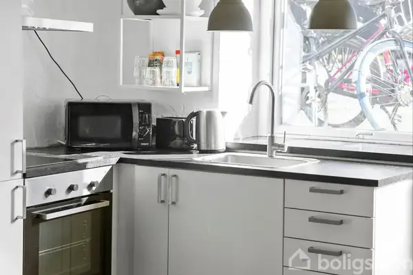 Et moderne køkken med hvide skabe, en ovn og mikrobølgeovn på bordpladen. En elkedel står ved siden af vasken under et vindue, hvor cykler kan ses udenfor.