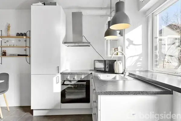 Et moderne køkken med hvide skabe og en sort bordplade. Der er en indbygget ovn, mikrobølgeovn, elkedel og emhætte. To lamper hænger over, og et vindue giver naturligt lysindfald.