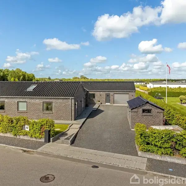 Et moderne hus med mørkt tag og murstensfacade står langs en stille vej. Foran huset er en indkørsel omgivet af grønne hække. Baggrunden viser åbent landskab og himmel.